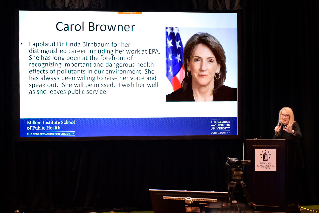 Lynn Goldman, M.D. standing at podium with Carol Browner, J.D. shown on a projection screen with a message of goodwill