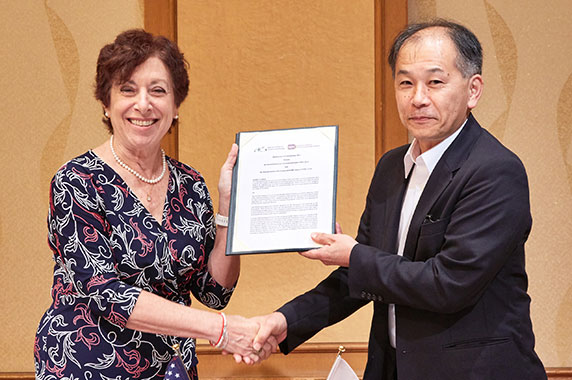 Birnbaum and Watanabe shake hands while holding sign MOU