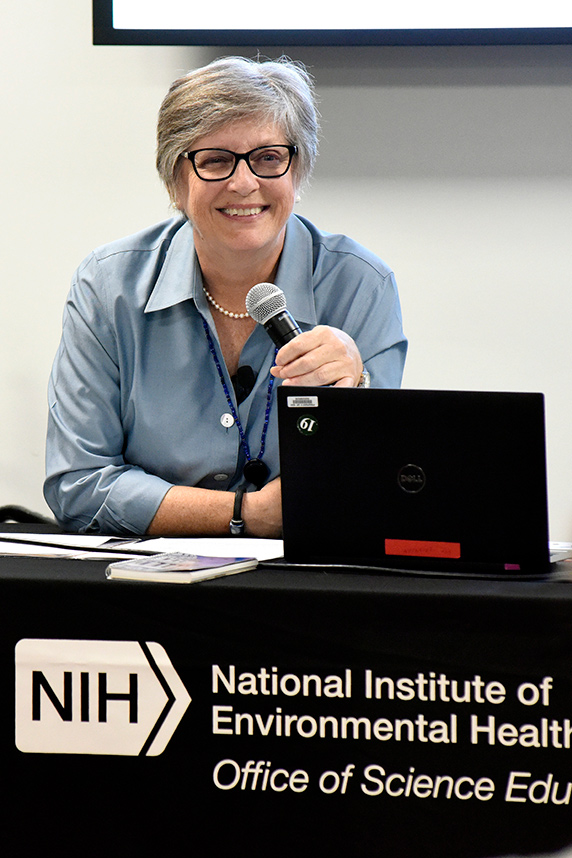 Stella Sieber holding microphone and sitting at table with NIH/NIEHS Office of Science Edu banner