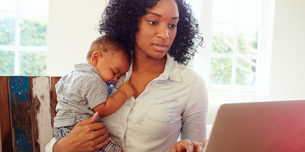 woman holding a sleeping baby while looking at a laptop