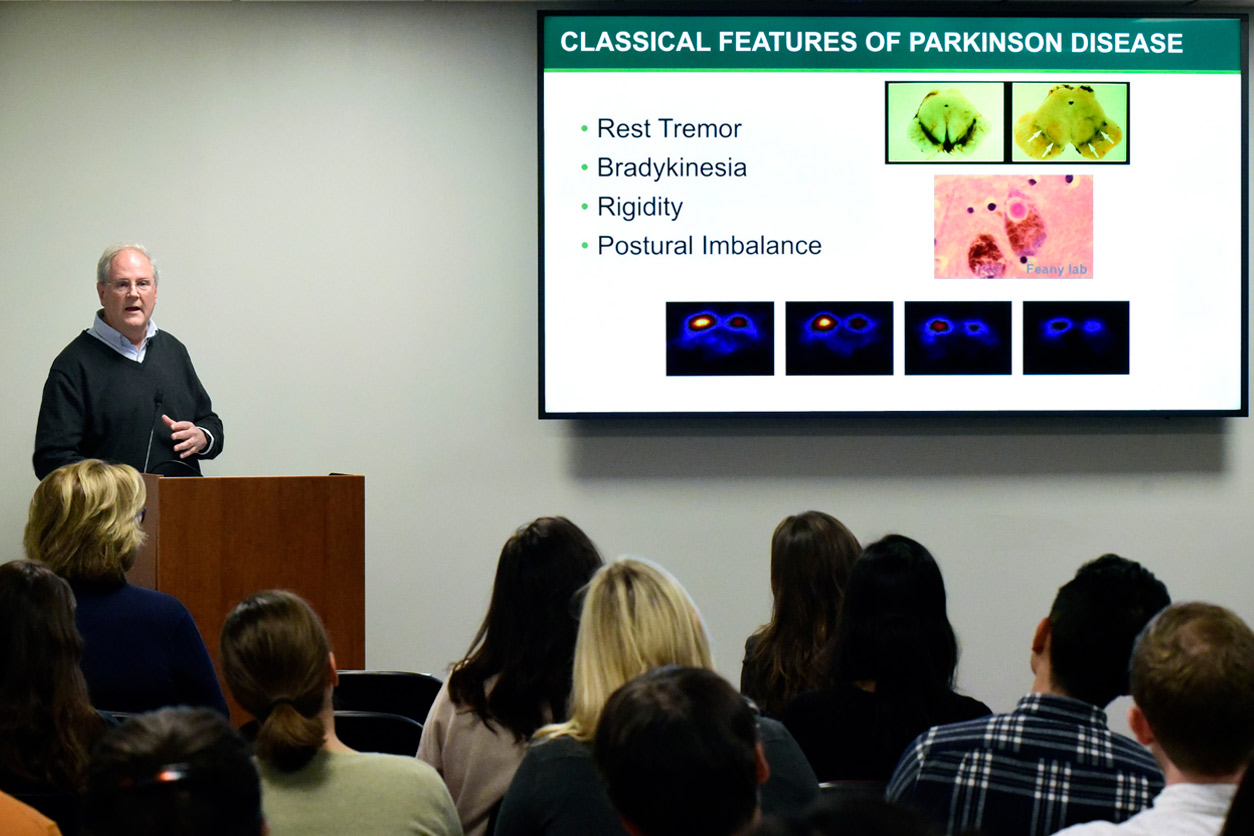 David Standaert, M.D., Ph.D. speaks to audience with Classical Features of Parkinson Disease on projection screen