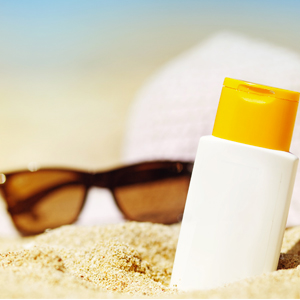 closeup of bottle of sunscreen slightly buried in sand with sunglasses and hat blurred in the background