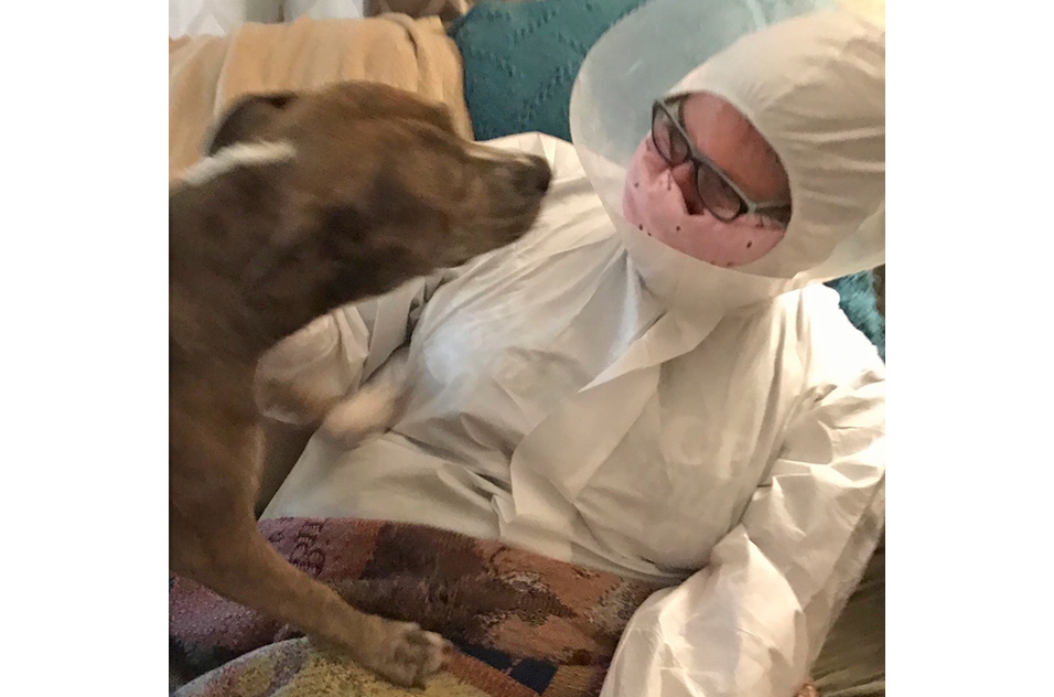 Susan Booker wears coveralls, a mask, and an Elizabethan cone as her dog steps on her lap