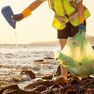 Woman in yellow safety vest picks up plastic garbage beside a body of water